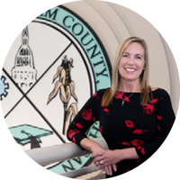 Barb Byrum, a white woman with blonde hair in a black dress with red flowers stands in front of the Ingham County MI seal
